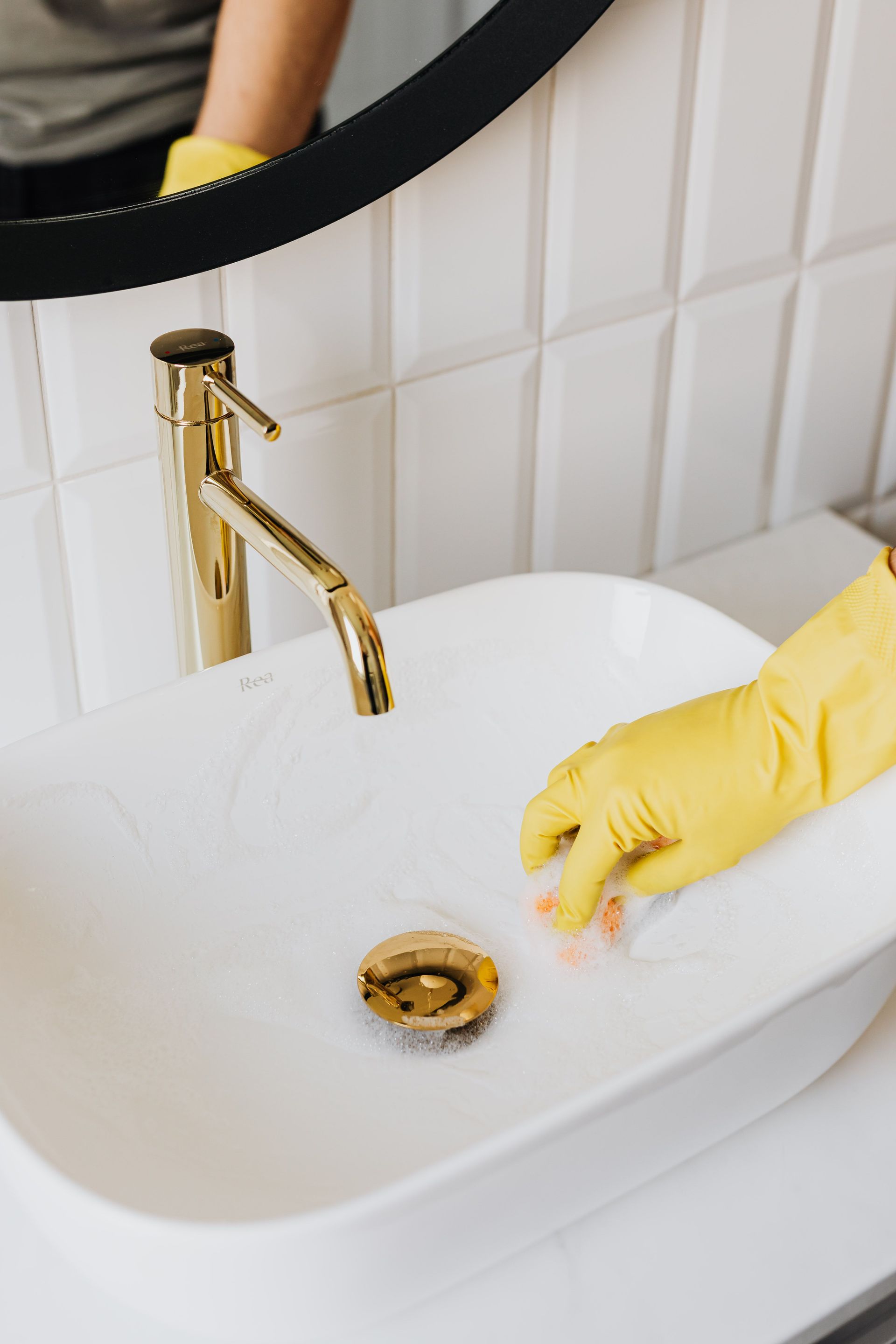 From above anonymous housekeeper in yellow rubber gloves wiping by sponge sink in bathroom